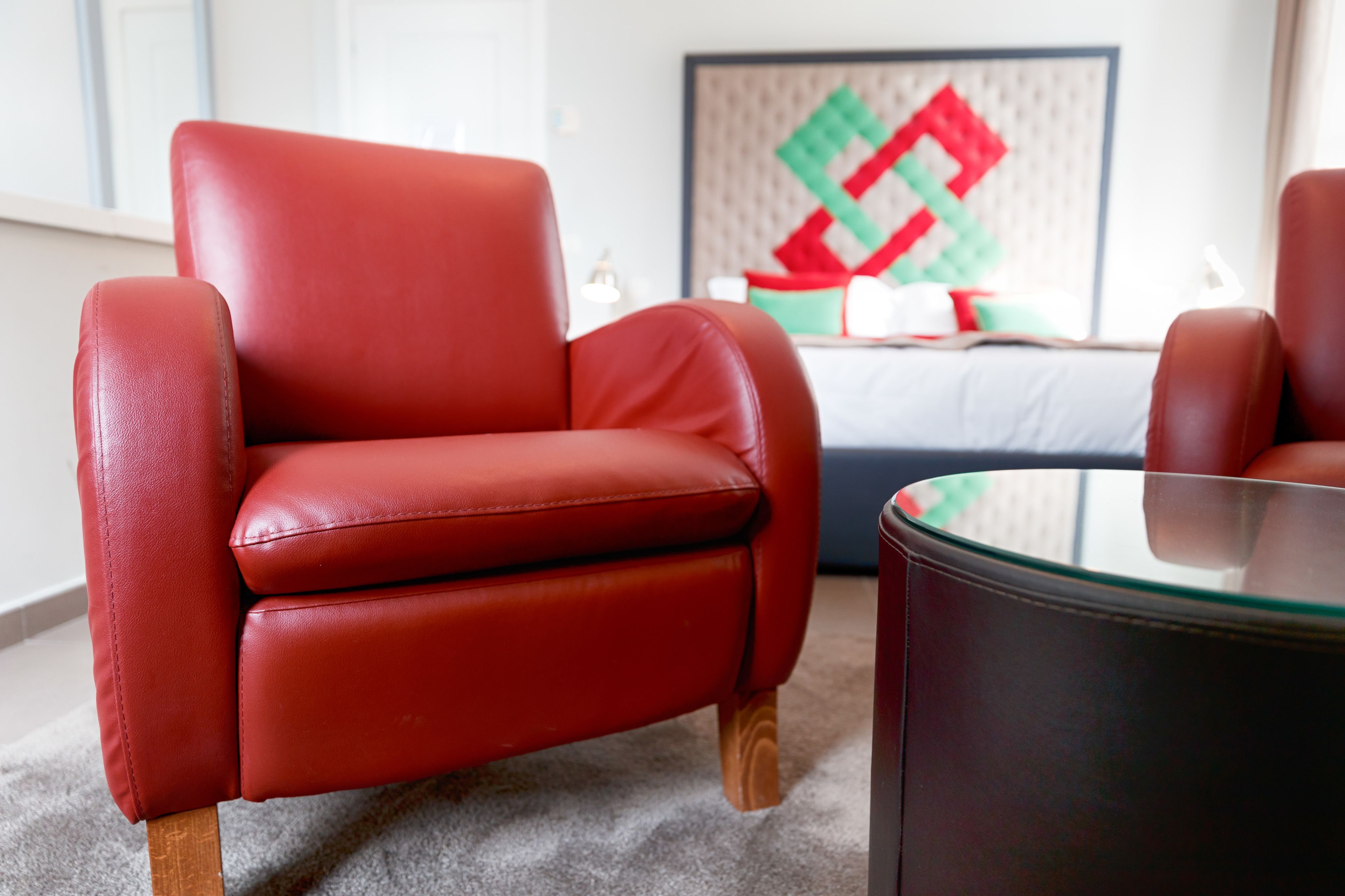 Un sillón rojo de cuero con patas de madera, una mesa de centro de vidrio y una cama con cabecera de estampado geométrico en una habitación de hotel.