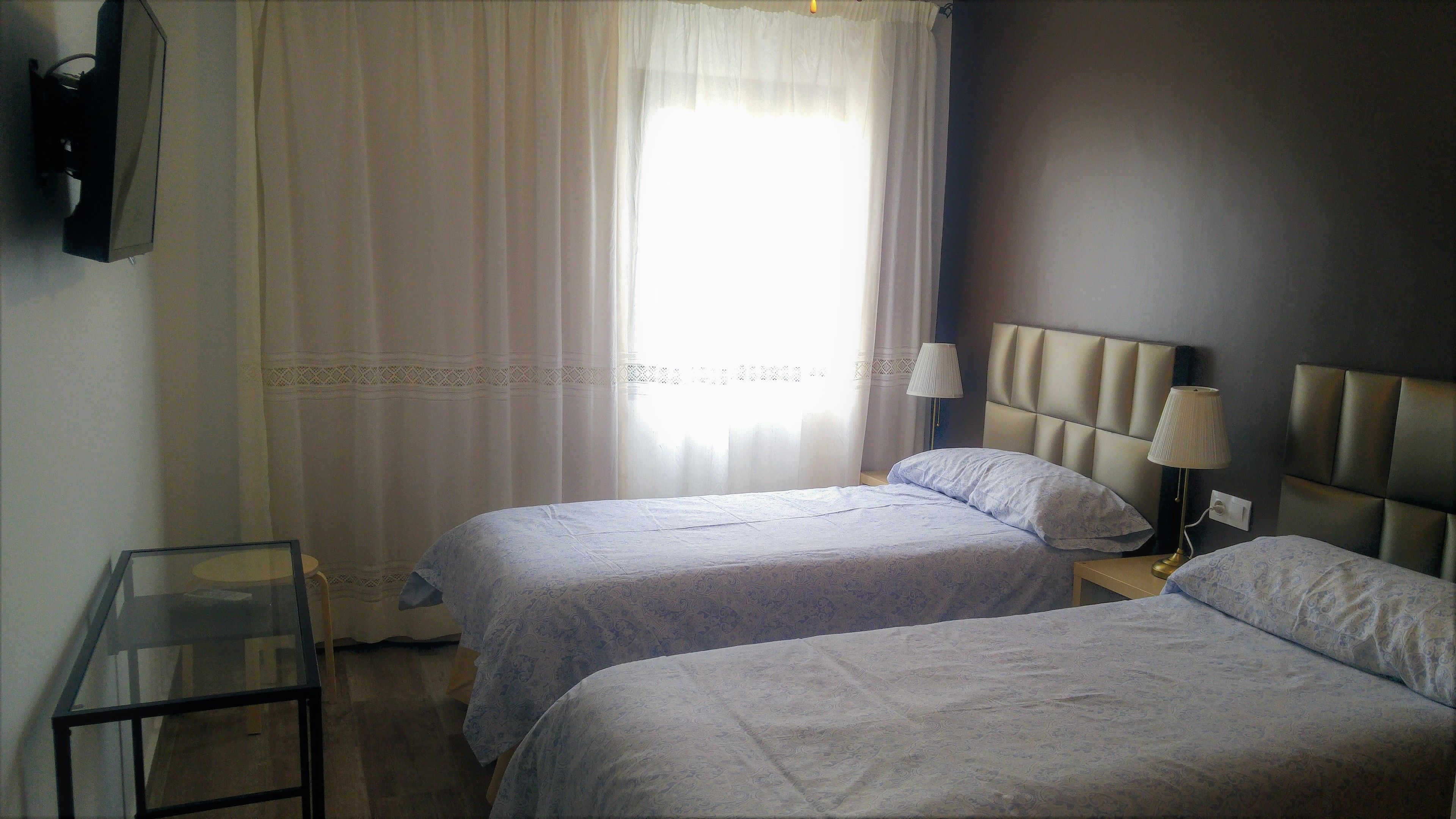 A hotel room with two beds, a bedside table, a lamp, and a window with sheer curtains.