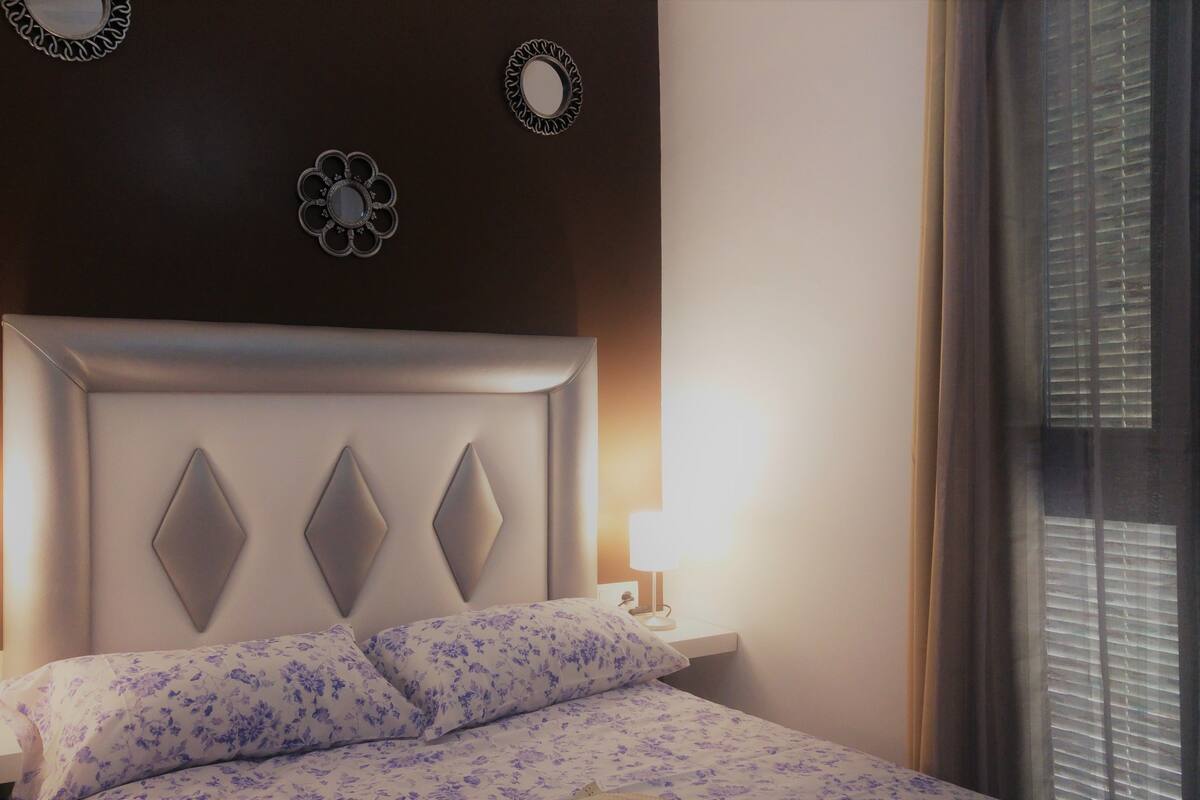 A neatly made bed with a floral-patterned bedspread, a headboard with diamond-shaped panels, and two bedside lamps.