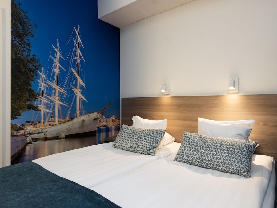 A modern bedroom with a large bed, two pillows, a wooden headboard, and a wall mural of a sailing ship.