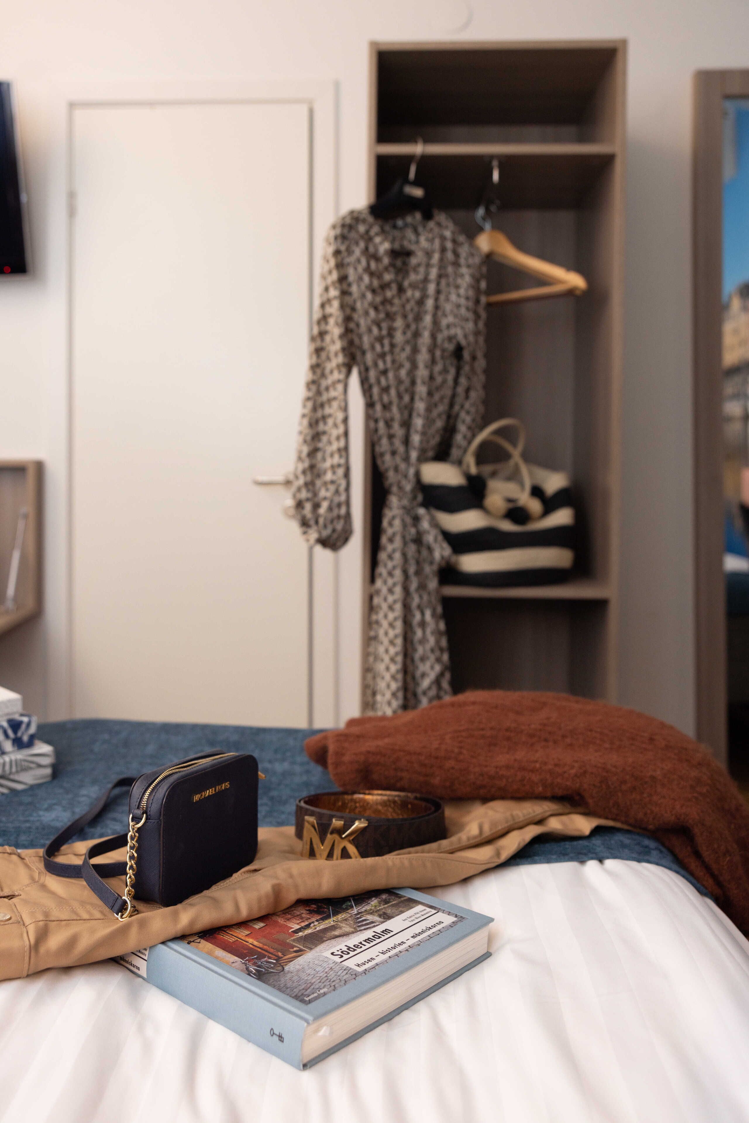 A bed with a book, a bag, and a pillow. A closet with clothes and shoes is in the background.