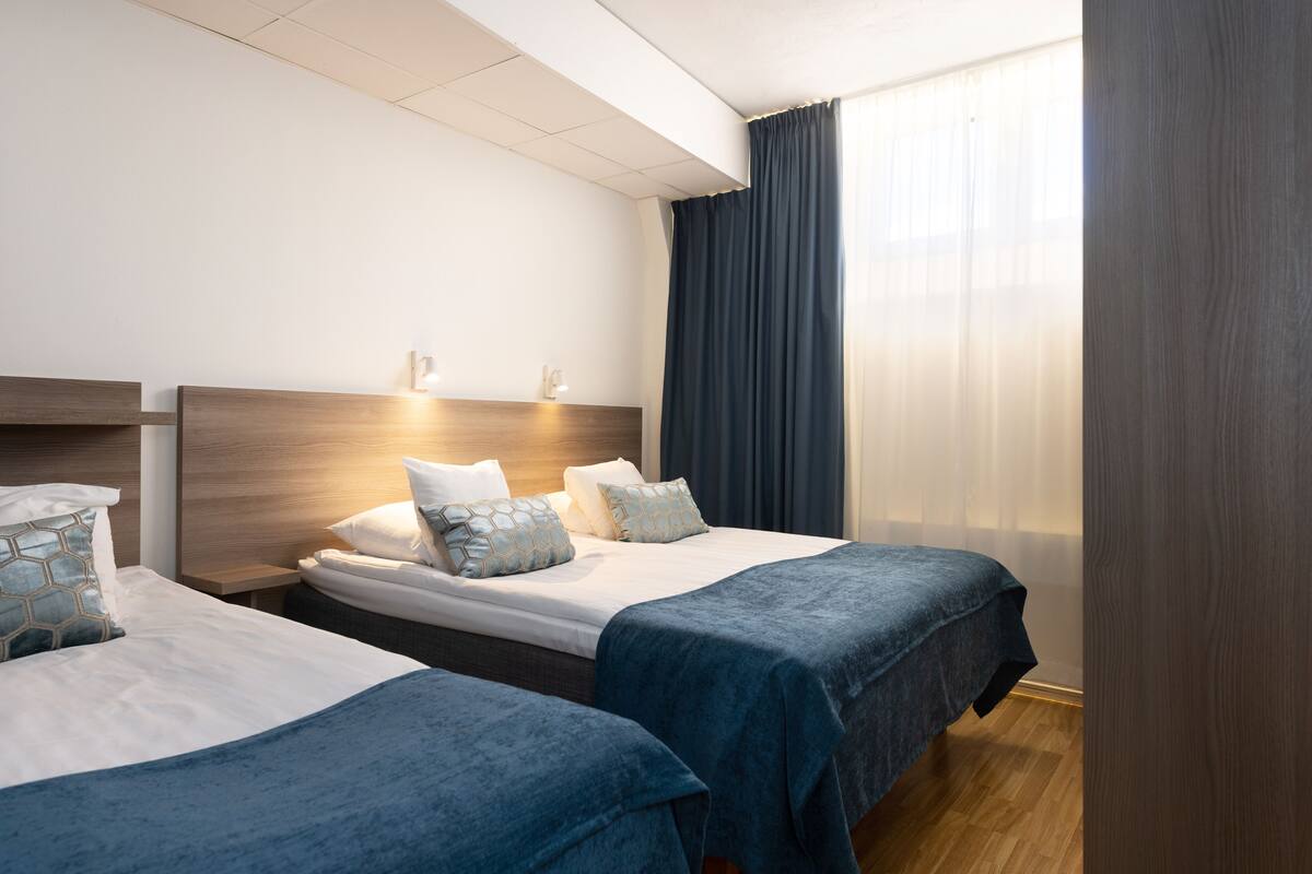 Habitación de hotel con dos camas, cabecera de madera, mesita de noche y ventana con cortinas.