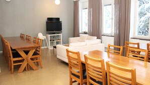 A room with a dining table, chairs, a sofa, a TV on a stand, and large windows with curtains.