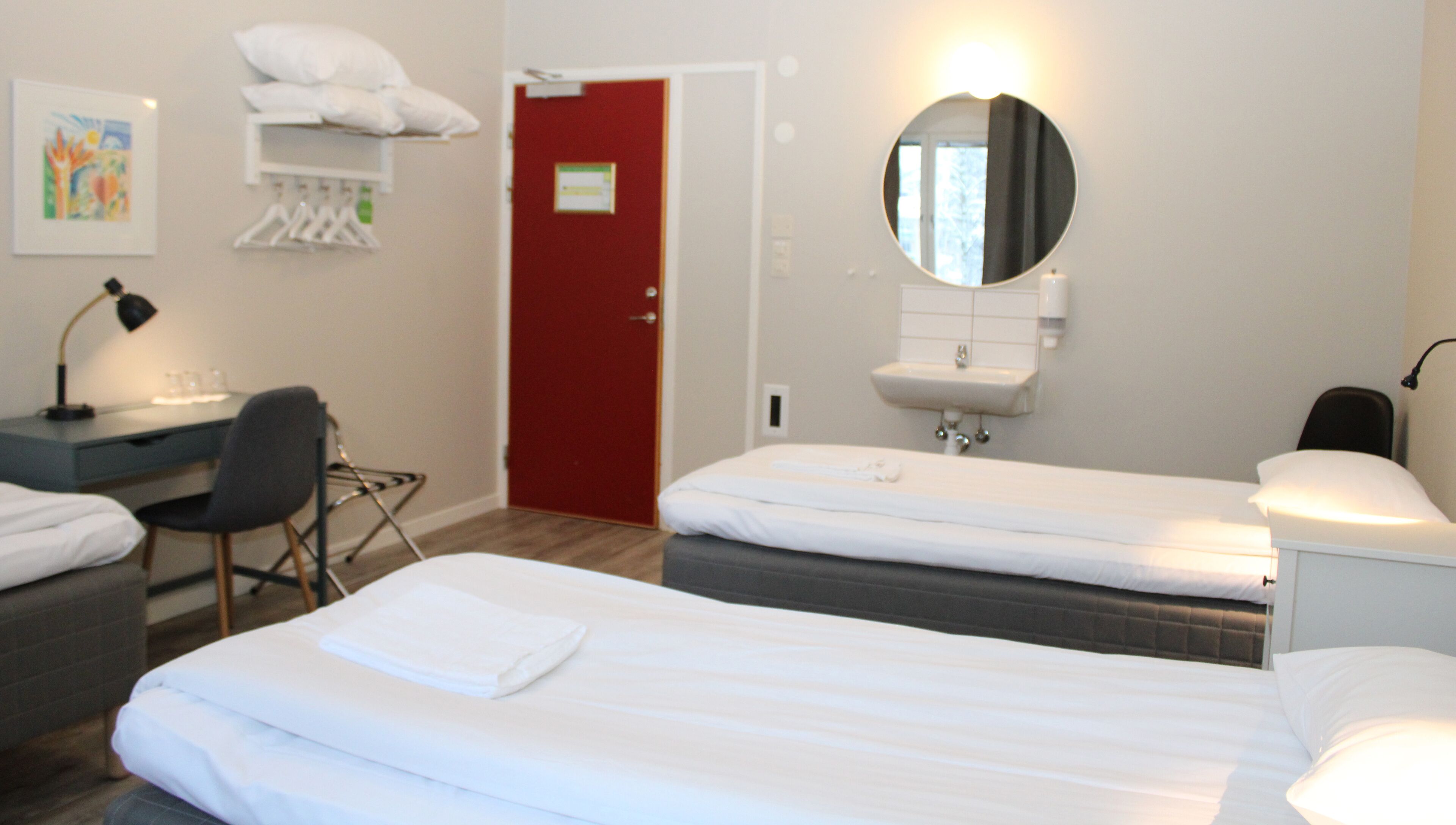 A hotel room with two beds, a desk, a chair, a red door, a mirror, and a wall-mounted shelf with towels.
