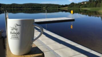 Una taza blanca con texto sobre una superficie de madera junto a un lago tranquilo con un muelle.