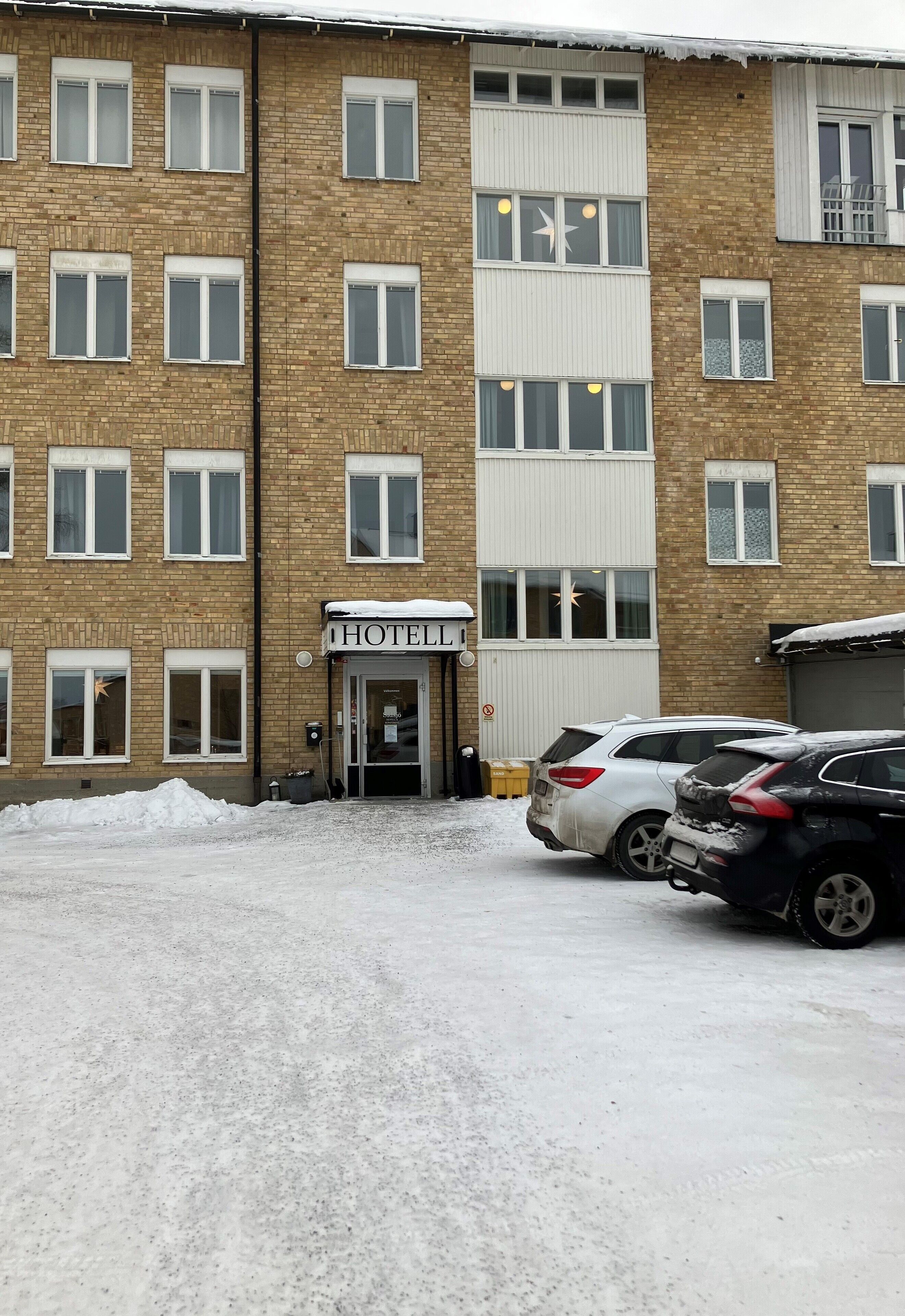 Um hotel de vários andares com fachada de tijolos, entrada coberta de neve e dois carros estacionados em frente.