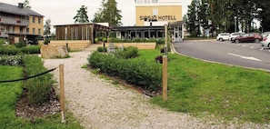 A hotel entrance with a gravel path leading to a wooden structure, a grassy area, and a road with parked cars.