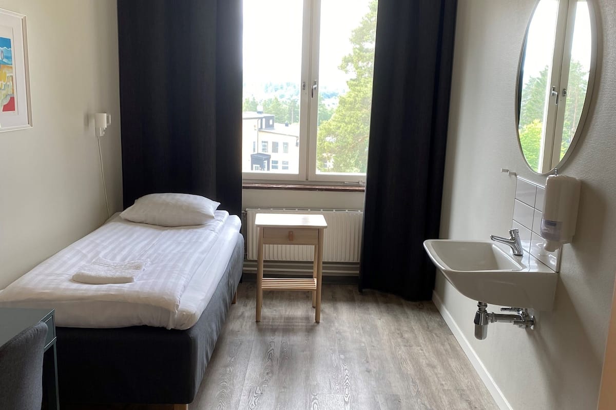 A small, tidy room with a bed, a desk, a chair, a window with curtains, and a bathroom with a sink and mirror.