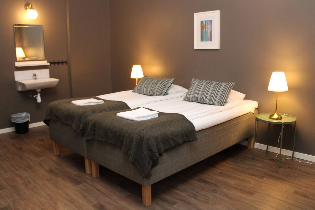 A hotel room with a double bed, two bedside lamps, a bedside table, a wall-mounted sink, and a trash bin.