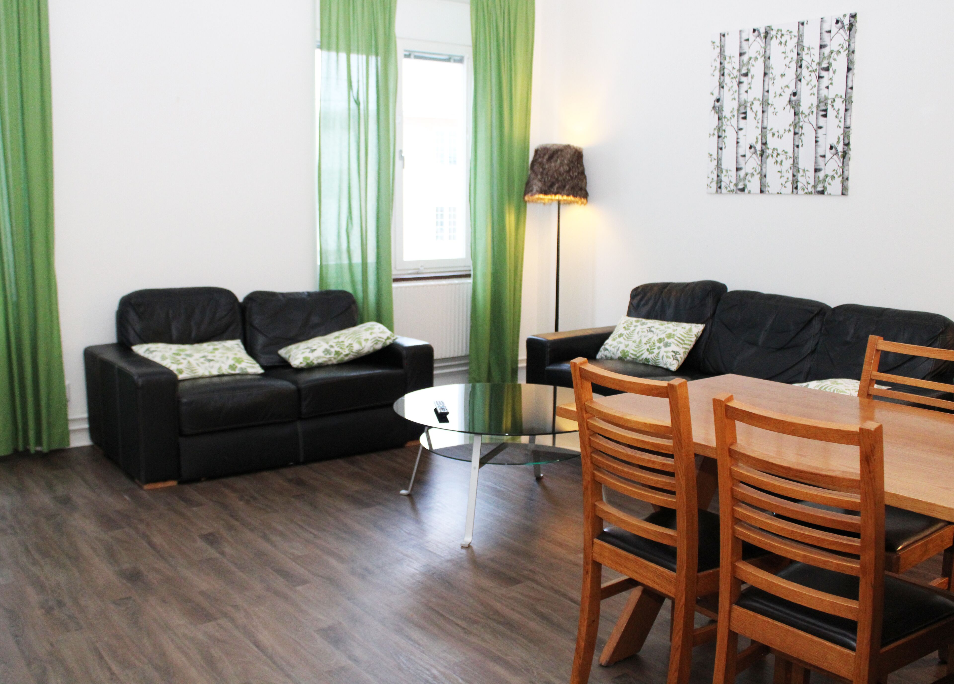 Una sala de estar con un sofá negro, una mesa de centro de vidrio, una mesa de comedor de madera con sillas y una lámpara de pie.