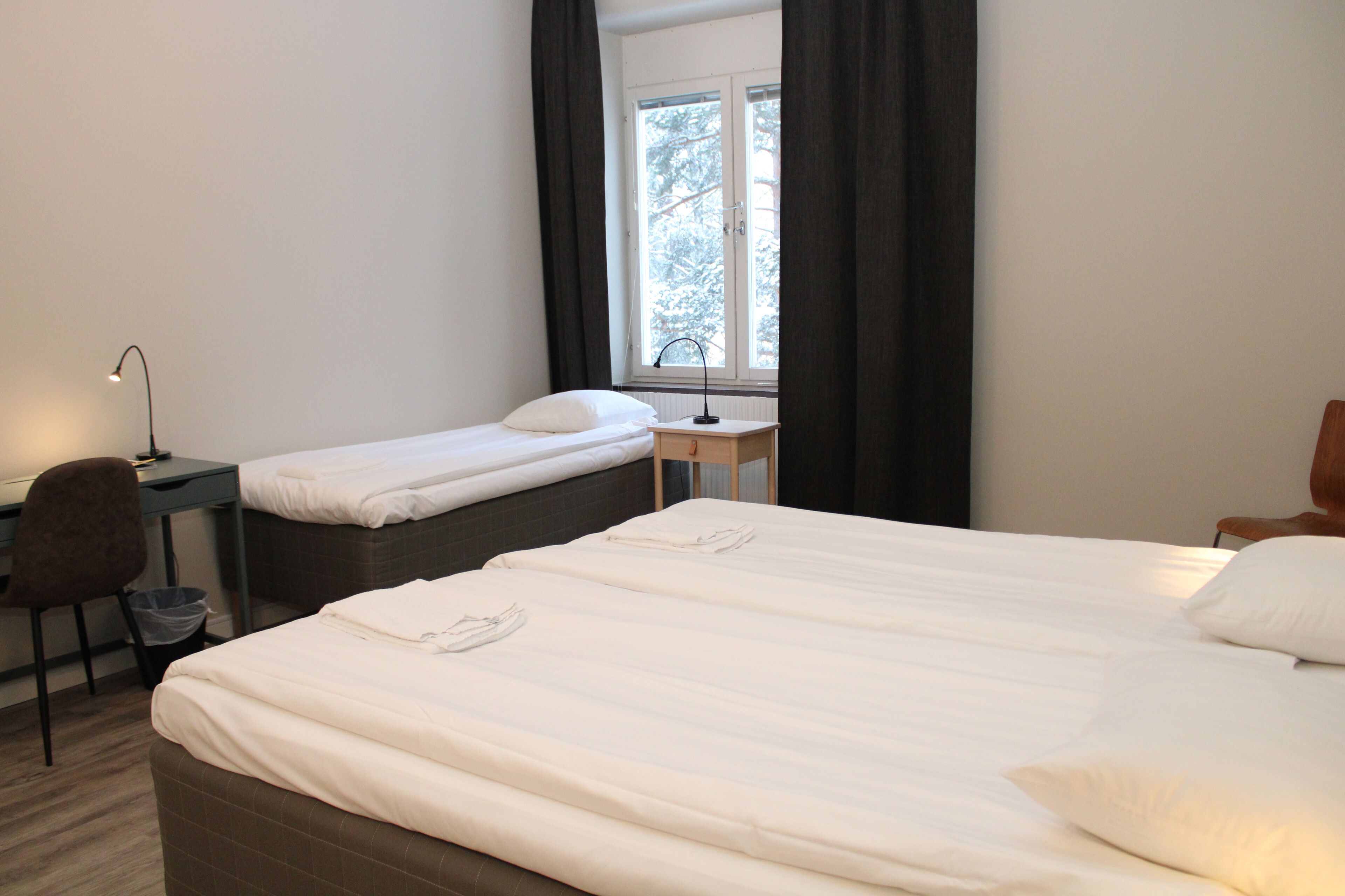 A hotel room with a large bed, a smaller bed, a desk, a chair, and a window with curtains.