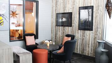 A cozy seating area with a round table, two chairs, and a small orange ottoman. There are framed pictures on the wall and a large window with a view.