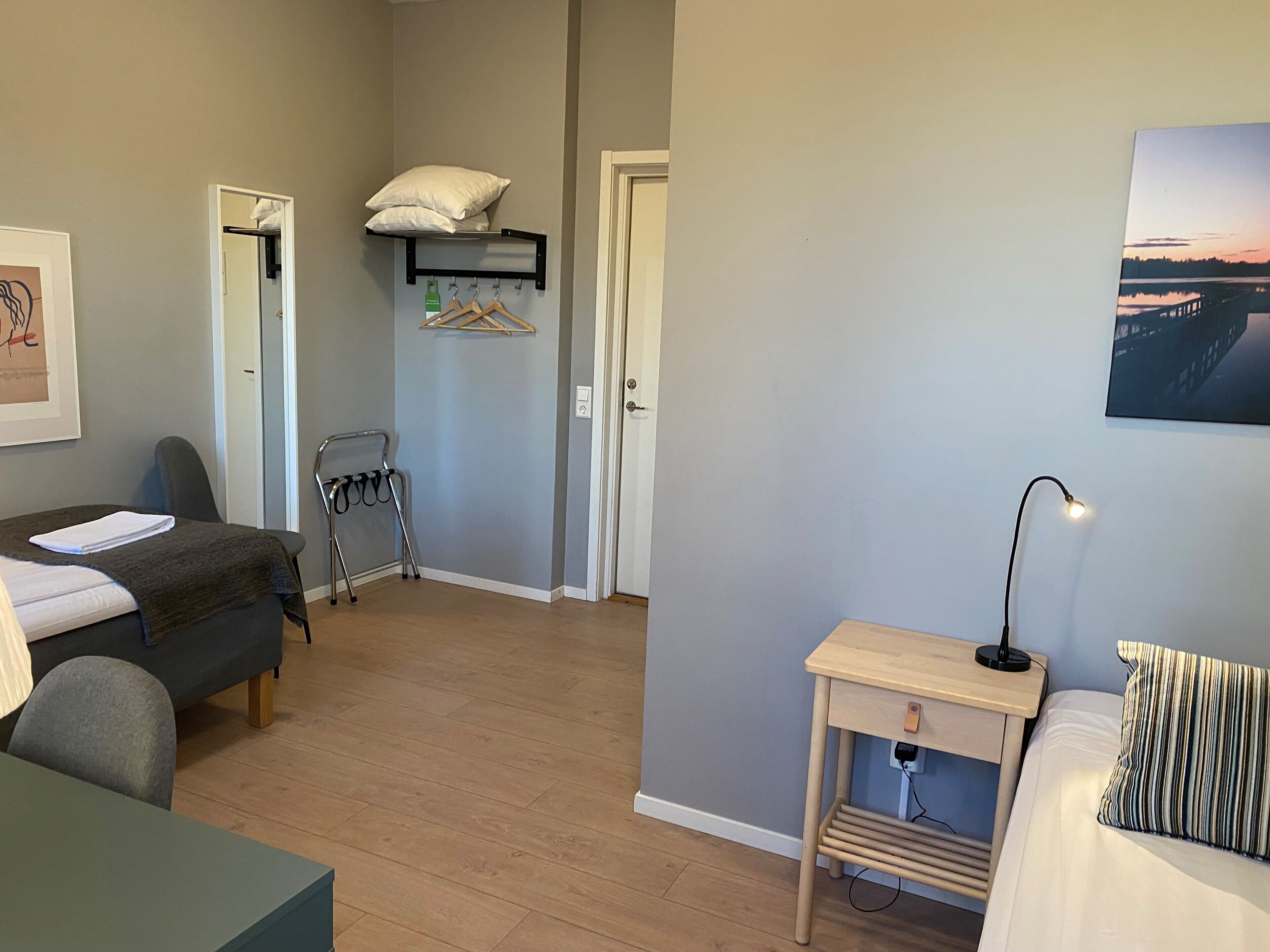 A compact room with a bed, a desk, a chair, a small table, and a hanging shelf with a pillow and clothes.