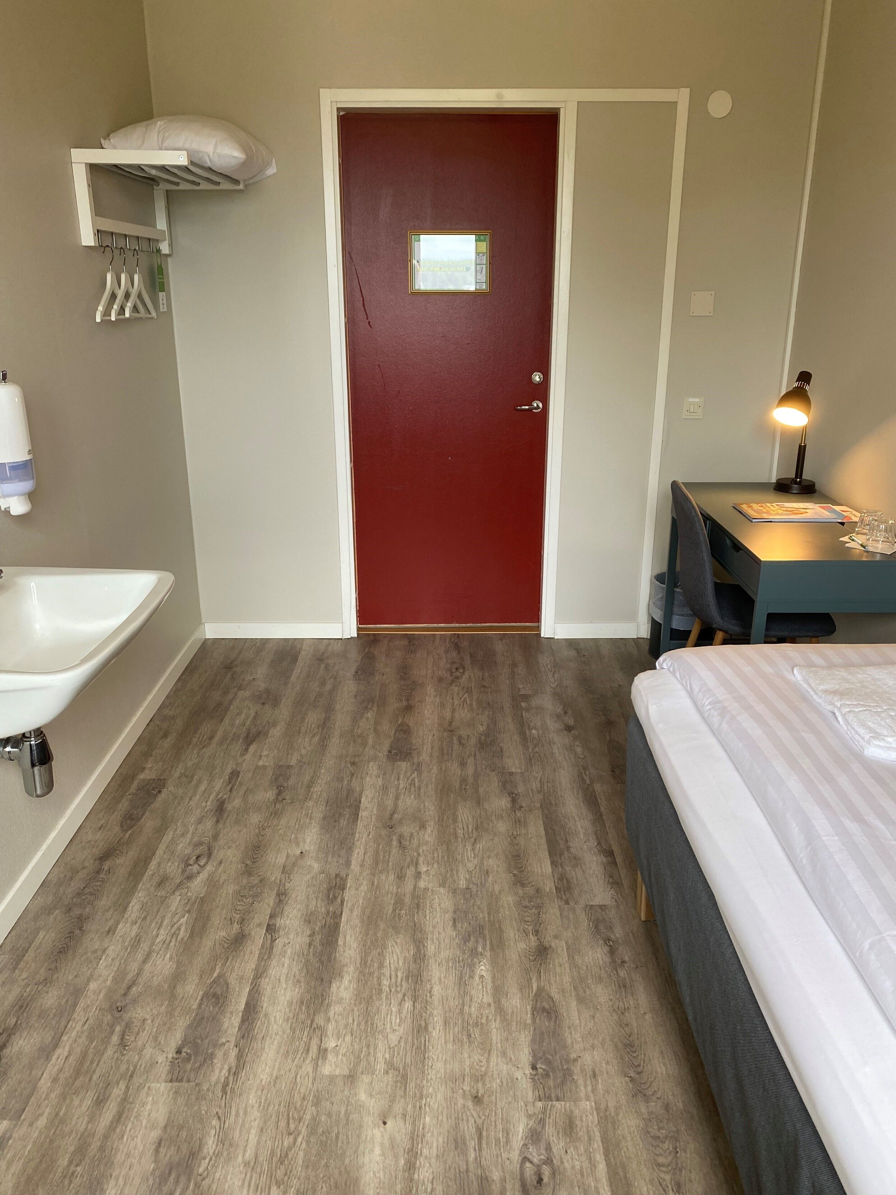 A hotel room with a wooden floor, a red door, a white sink, a bedside table with a lamp, and a bed with a striped bedspread.