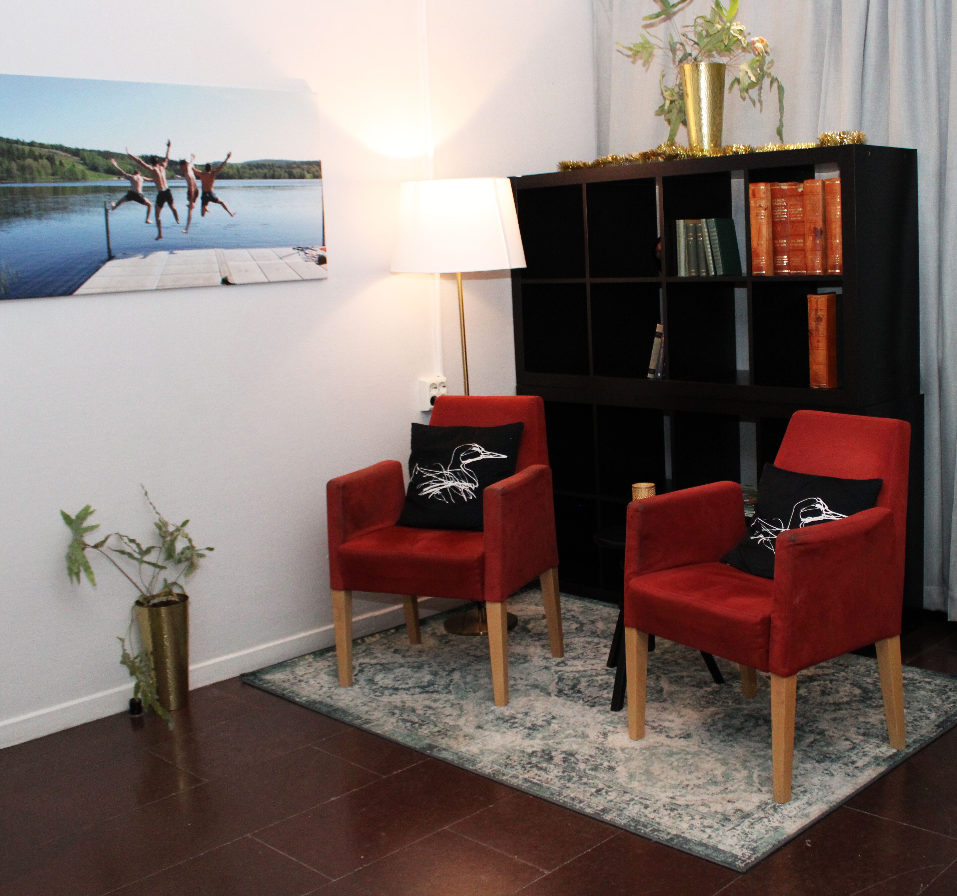 A living room with two red armchairs, a black bookshelf, a white wall, and a painting of a lake scene.