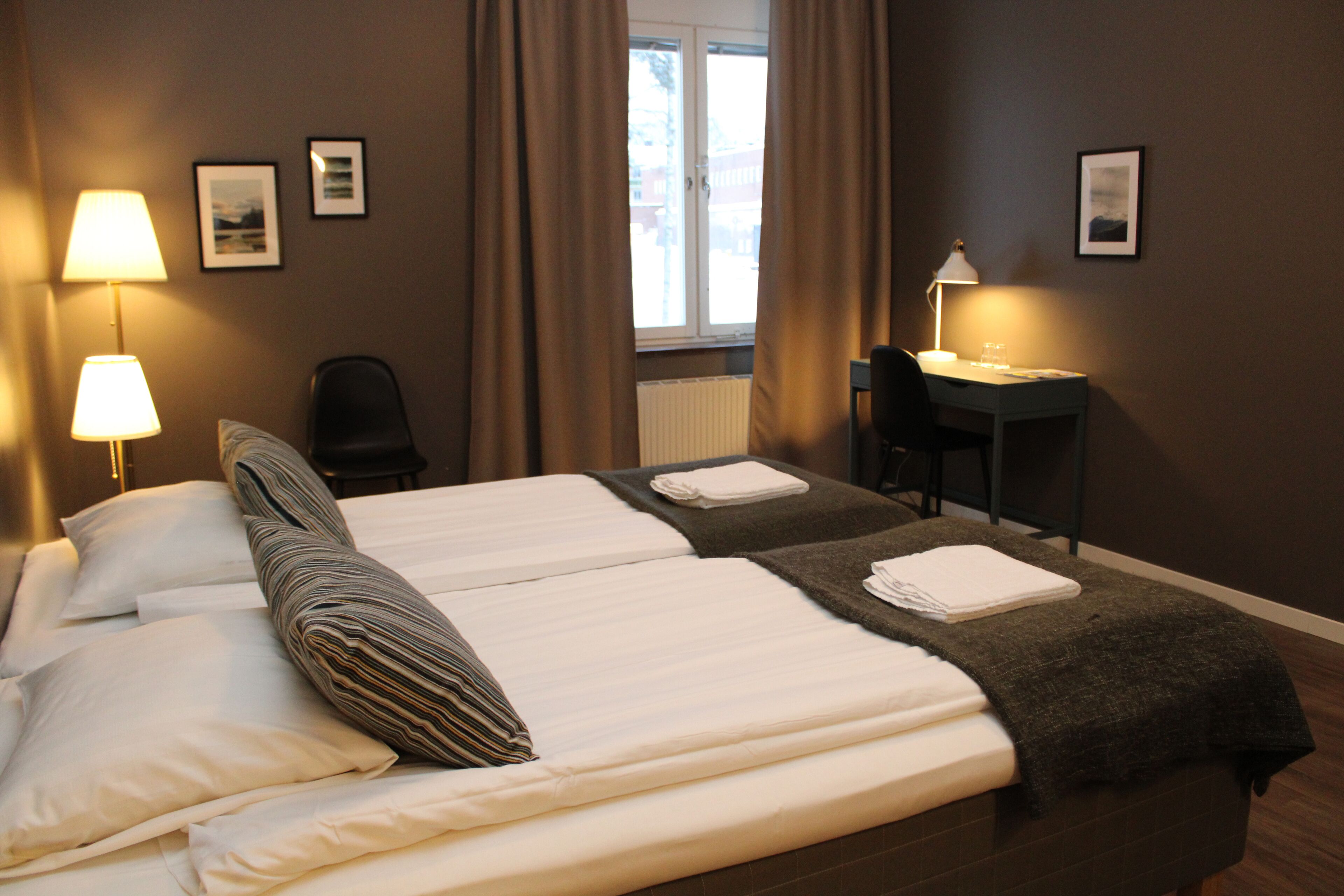 Habitación de hotel con dos camas, un escritorio, una silla y un ventanal con cortinas.