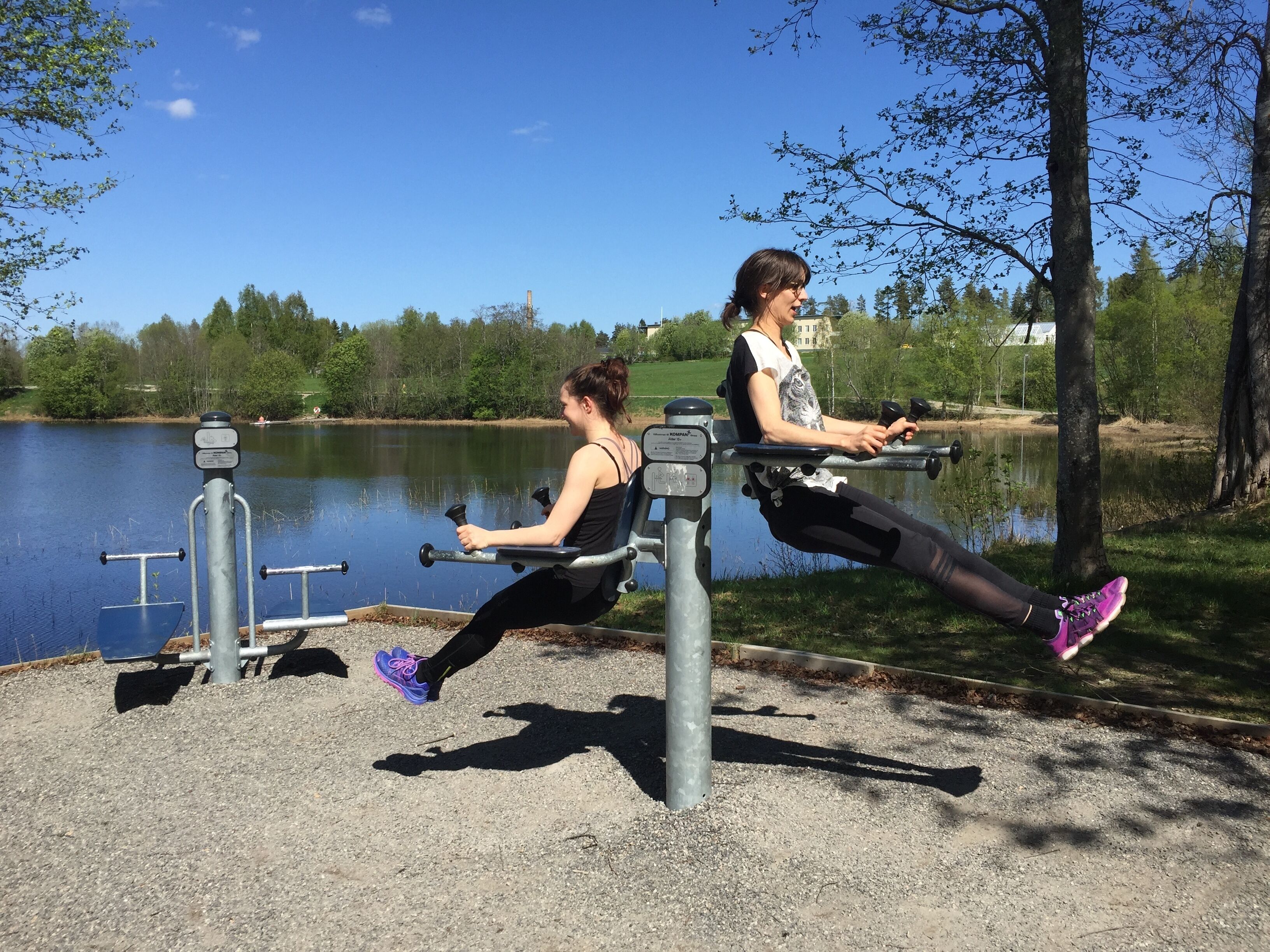 Two individuals exercising on outdoor stationary bikes by a lake with trees and a building in the background.