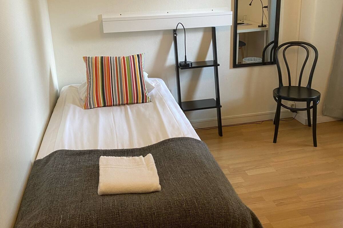 A single bed with a striped pillow, a bedside table with a lamp, a chair, and a framed artwork on the wall.