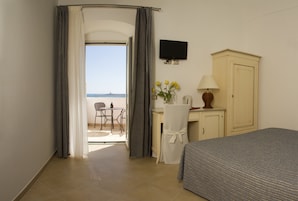 Quarto de hotel com cama, escrivaninha com cadeira, televisão e vista para o mar.