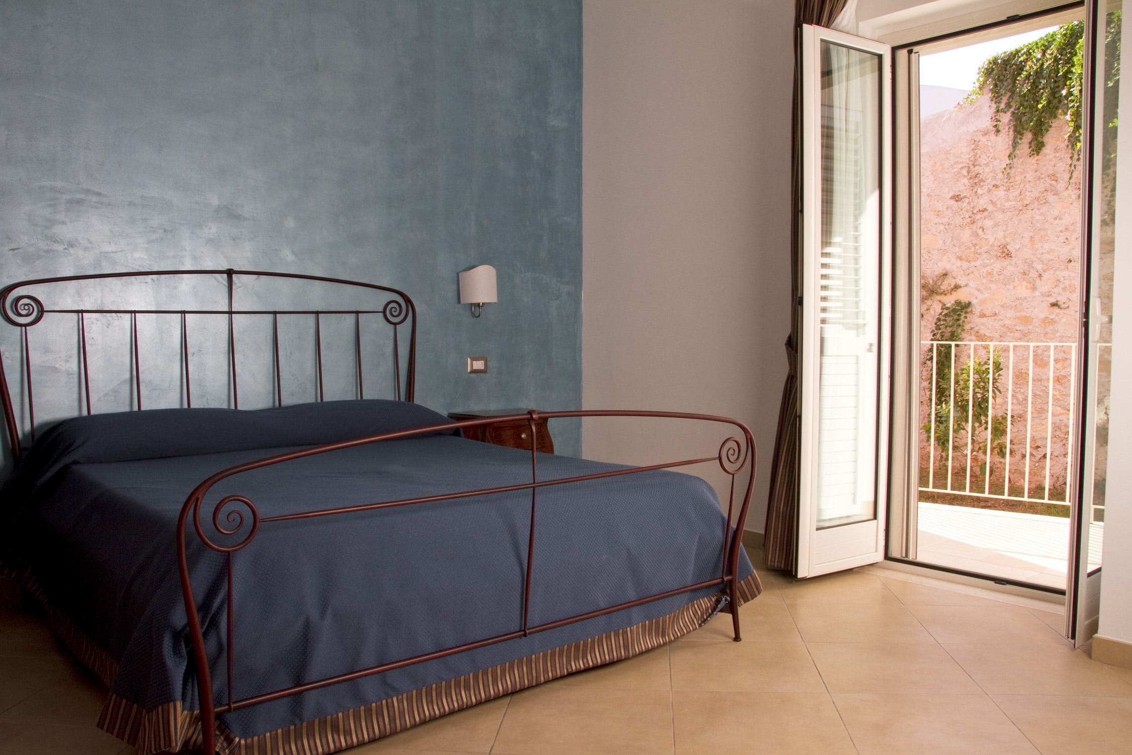 Une chambre à coucher avec un cadre de lit en métal, une couvre-lit bleue, une lampe de chevet et une porte-fenêtre coulissante donnant sur un espace extérieur.