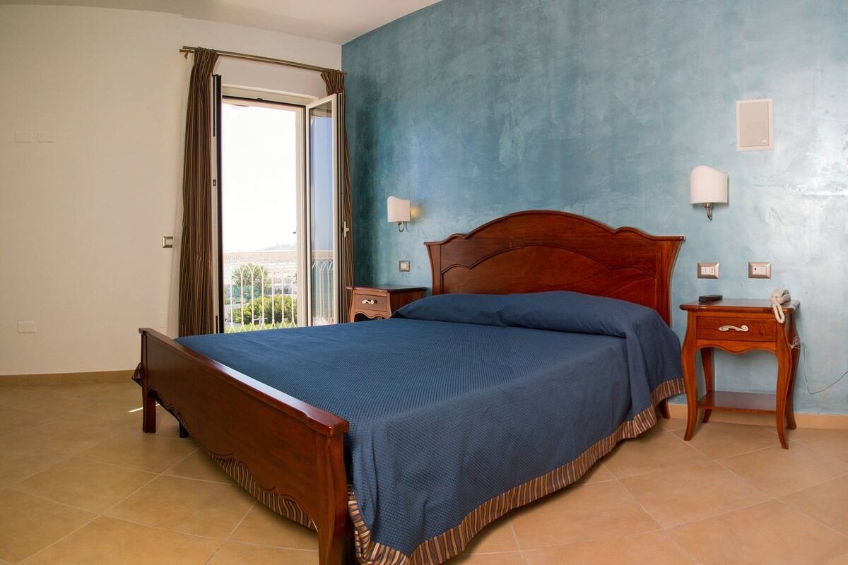 A bedroom with a wooden bed, a nightstand, a lamp, and a view through a window with curtains.