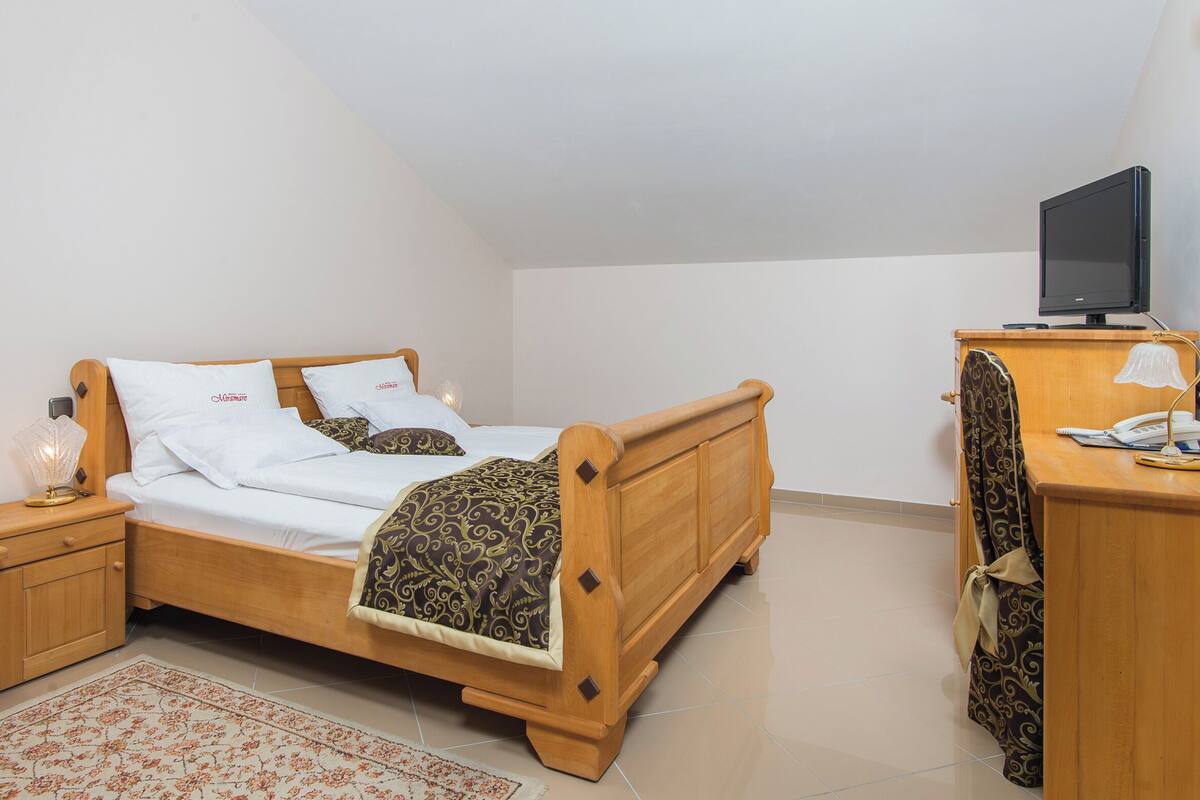 A bedroom with a wooden bed, a bedside table with a lamp, a TV on a wooden cabinet, and a patterned rug.
