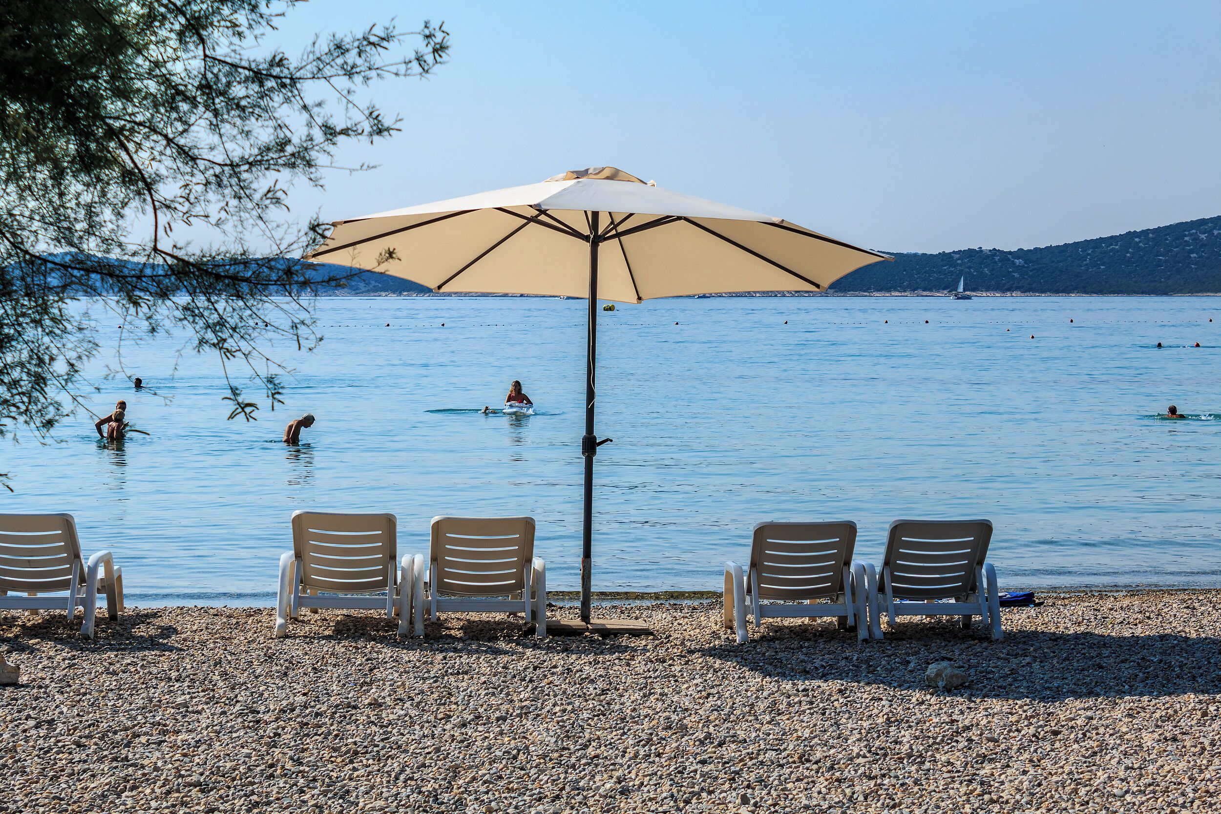 Ein Kiesstrand mit vier Liegestühlen und einem großen Sonnenschirm.