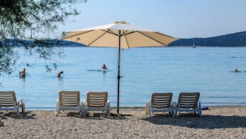 Ein Kiesstrand mit vier Liegestühlen und einem großen Sonnenschirm.
