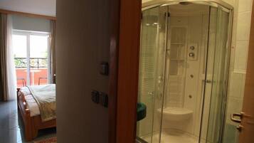 A hotel room with a glass-enclosed shower, a bed with a white duvet, and a view of a balcony with outdoor furniture.