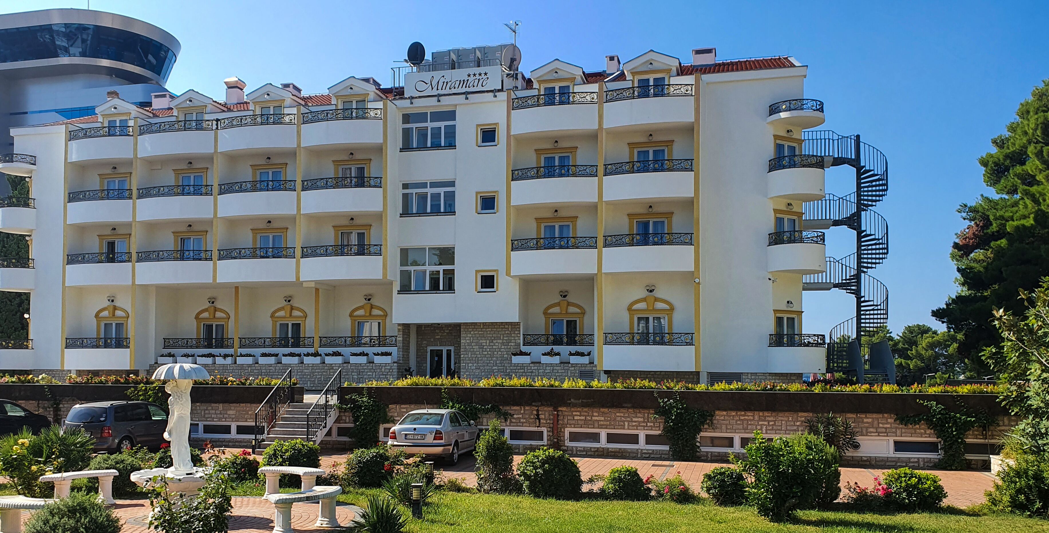 A multi-story hotel with balconies, a central entrance, and a garden area with a statue.