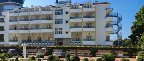 A multi-story hotel with balconies, a central entrance, and a garden area with a statue.