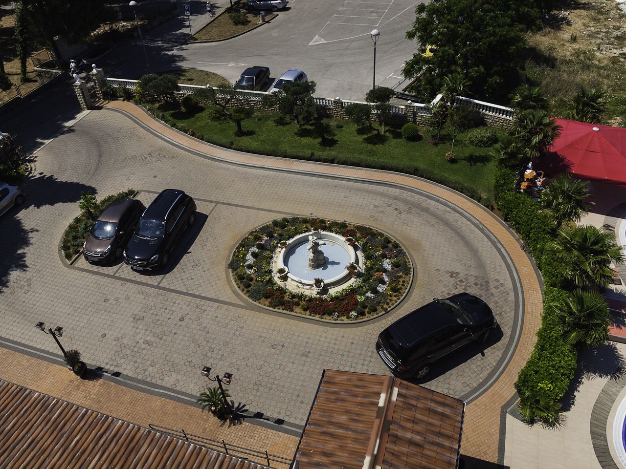 An aerial view of a parking area with a central circular fountain, surrounded by greenery and a few parked cars.
