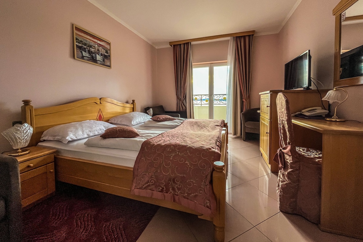 A hotel room with a wooden bed, a desk with a computer, a chair, a TV, and a balcony with curtains.