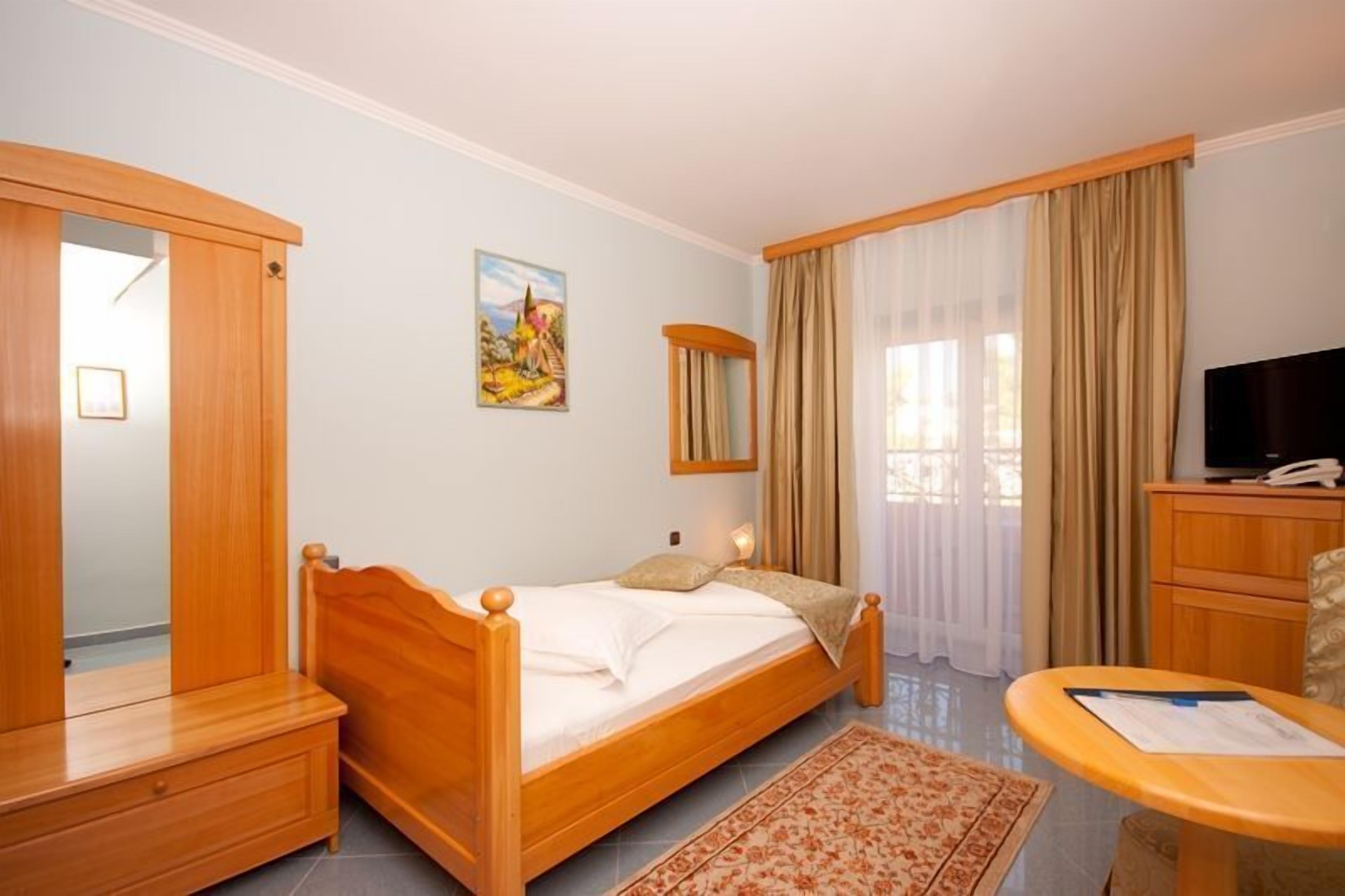 A bedroom with a wooden bed, a desk, a chair, a television, and a painting on the wall.