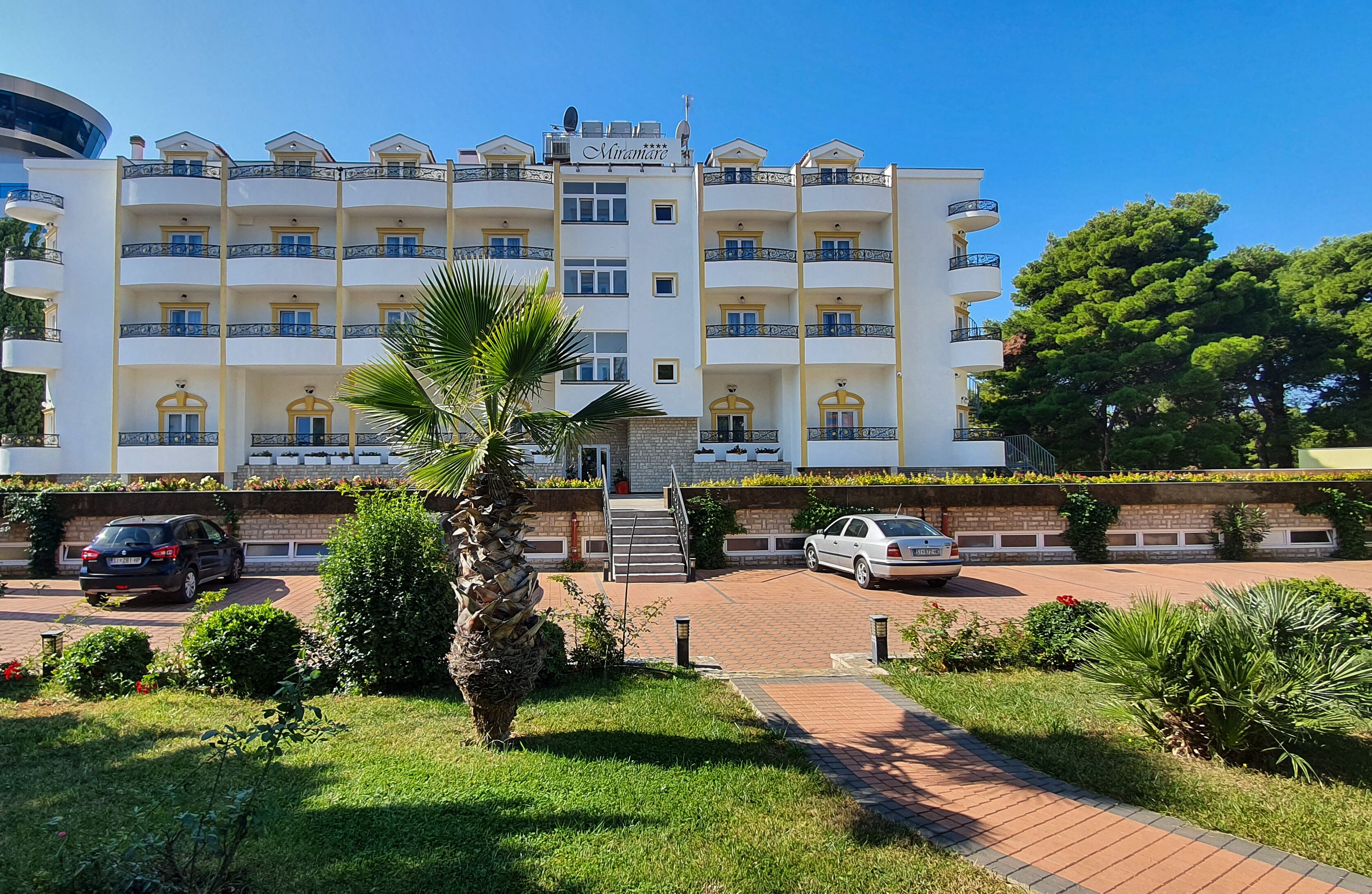 A multi-story hotel with a central entrance, balconies, and a well-maintained garden.