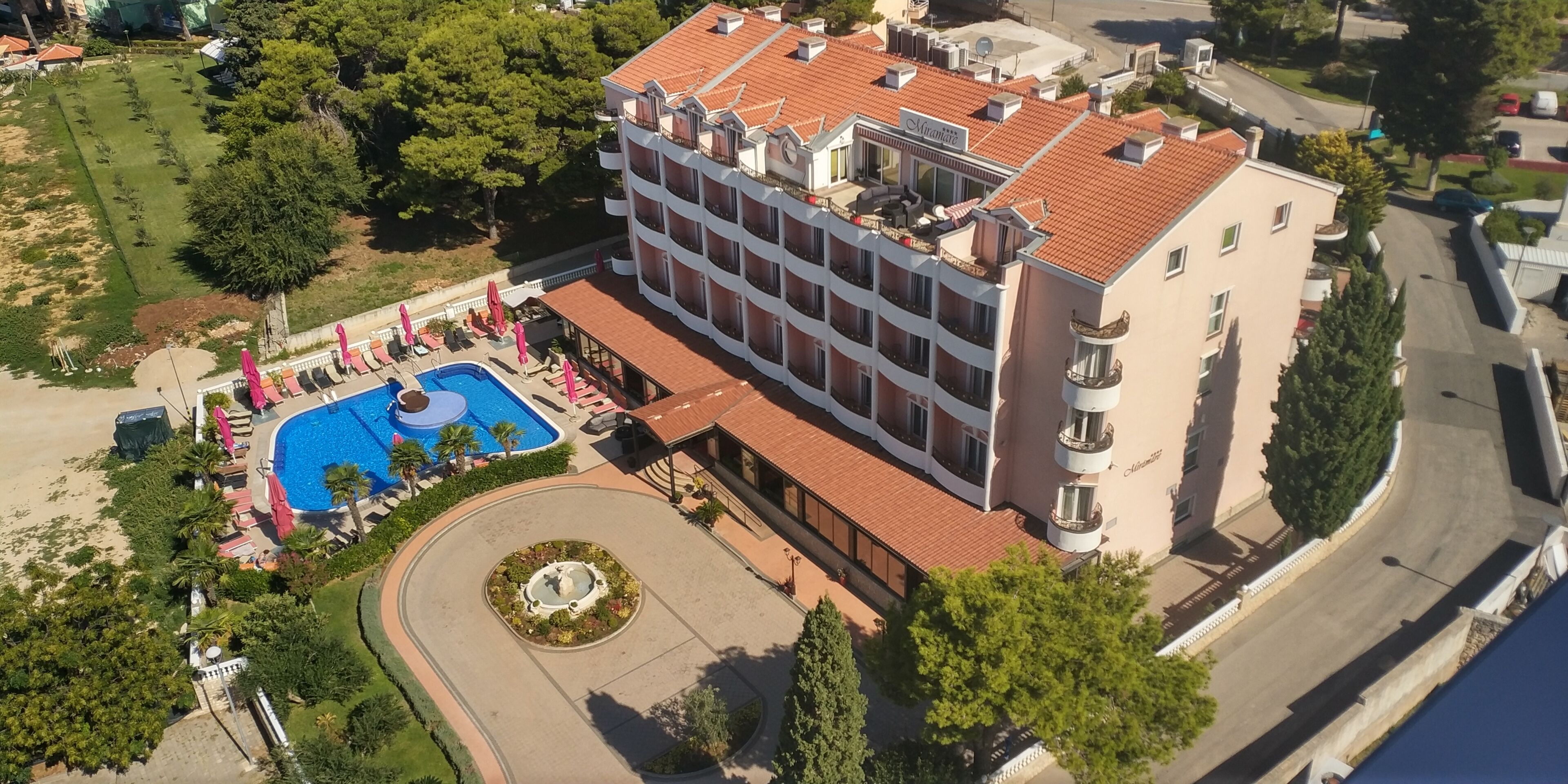 An aerial view of a hotel with a swimming pool, a circular fountain, and a landscaped garden.