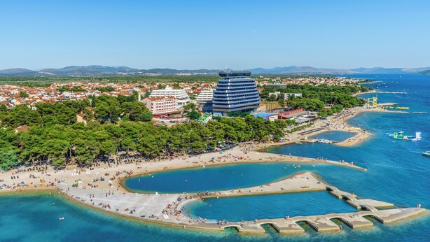 An aerial view of a coastal city with a prominent modern building, a beach, and clear blue waters. - Hotel Miramare (Vodice)