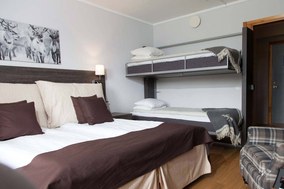 A bedroom with a bunk bed, a single bed, a headboard, a wall-mounted light, a painting, a door, and a small table.