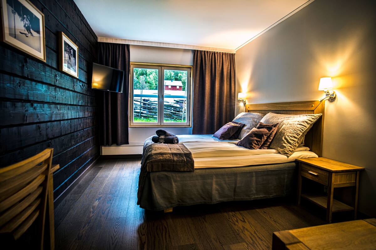 A bedroom with a bed, wooden floor, a window with curtains, a TV mounted on the wall, and a nightstand.