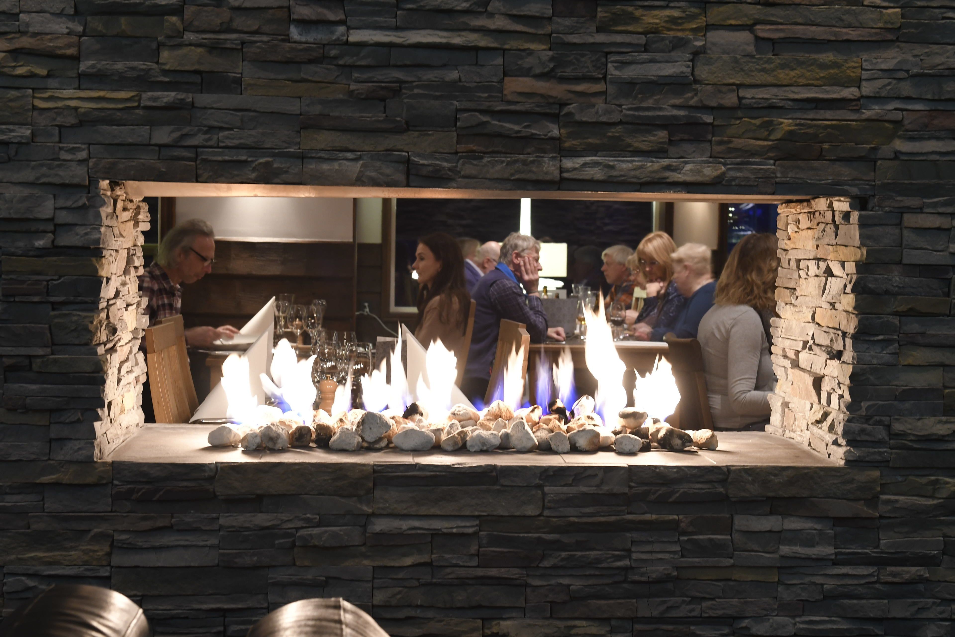 A stone fireplace with a fire, surrounded by seated guests at a bar area.