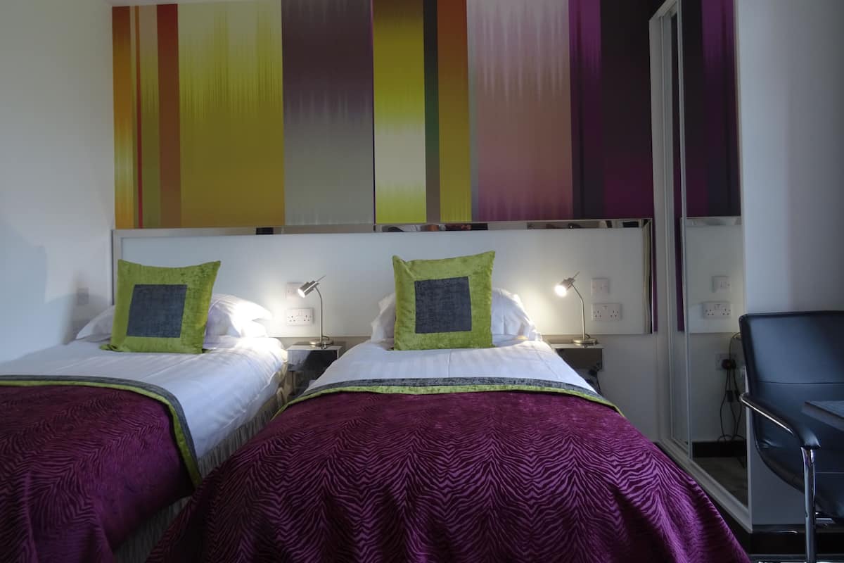 Quarto de hotel com duas camas, cada uma com almofada verde e manta roxa. Há uma mesa com luminária e uma cadeira.