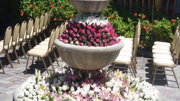 A tiered stone fountain with pink and white flowers, surrounded by chairs and greenery.