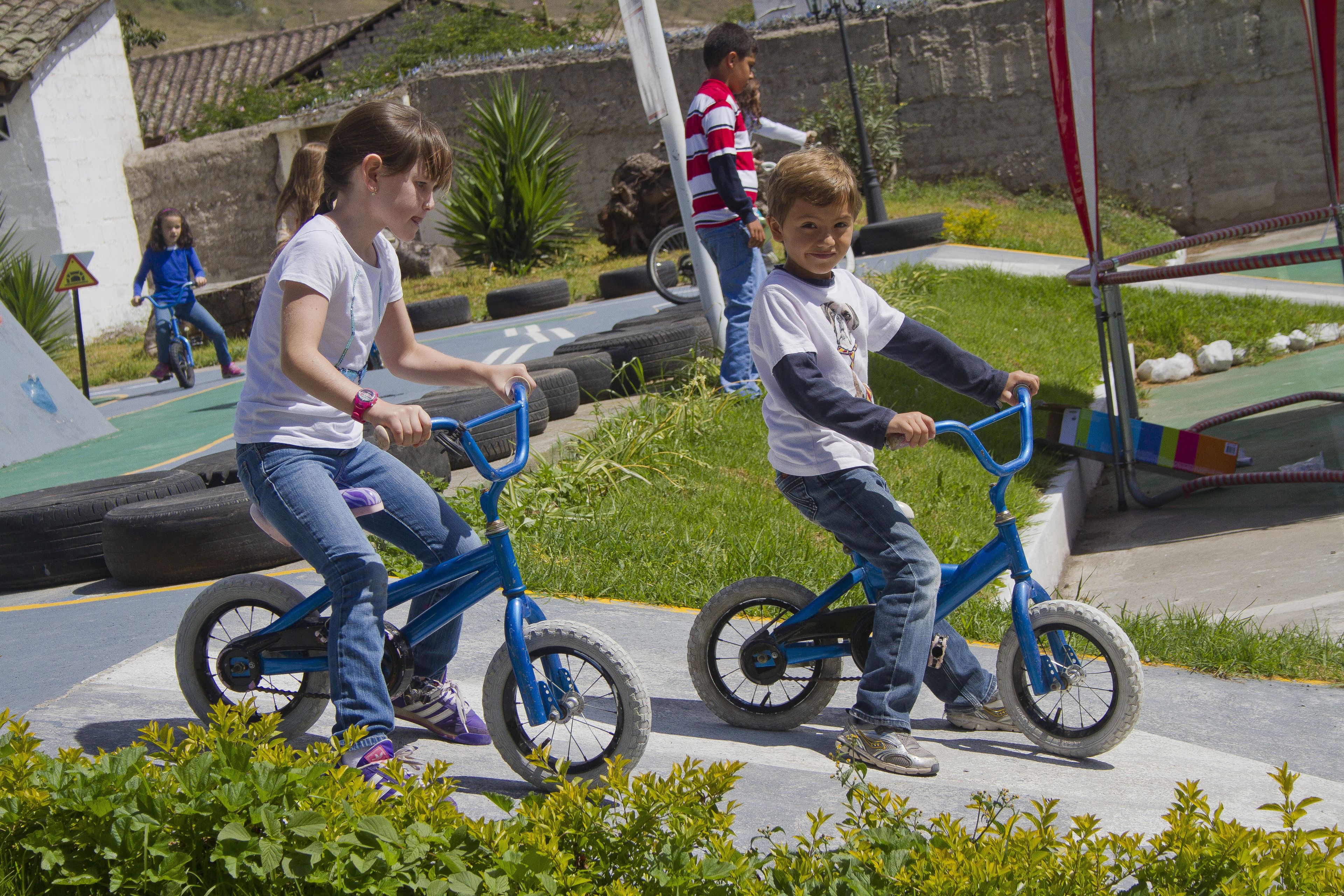 Children riding bicycles on a paved path with greenery and a playground in the background.