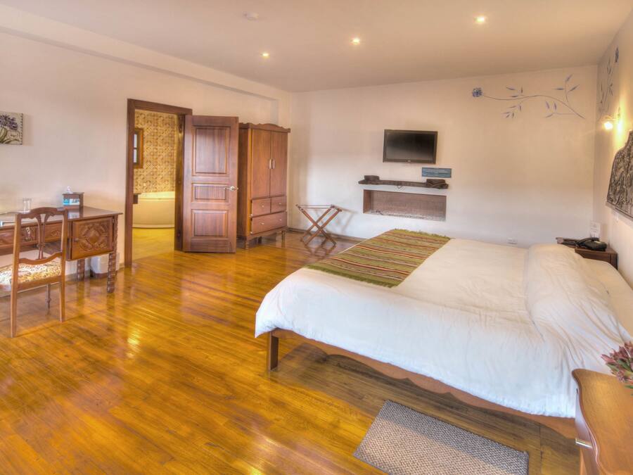 Quarto com cama de madeira, escrivaninha com cadeira, televisão fixada na parede e porta que dá para outro espaço.