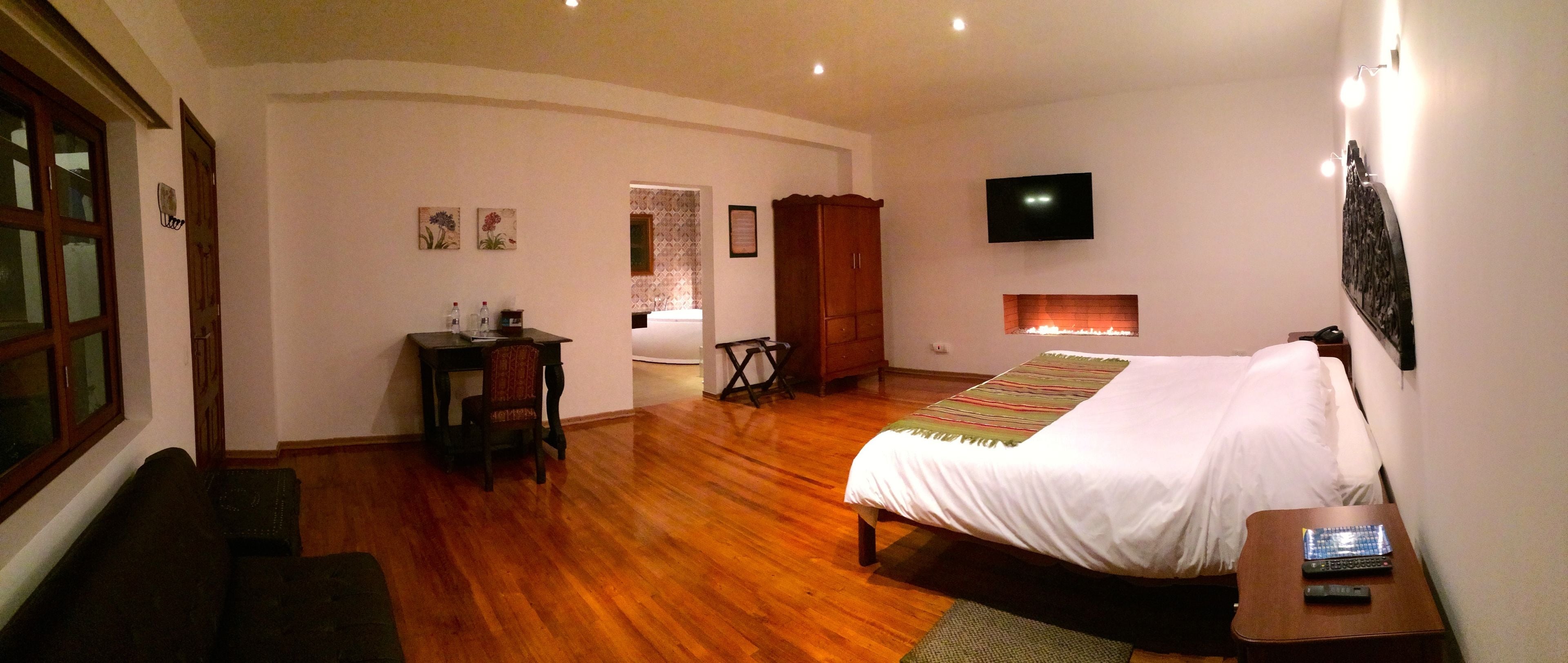 Ein Hotelzimmer mit Holzboden, einem Bett, einem Schreibtisch mit Stuhl, einem an der Wand montierten Fernseher und einem Fenster mit Vorhängen.