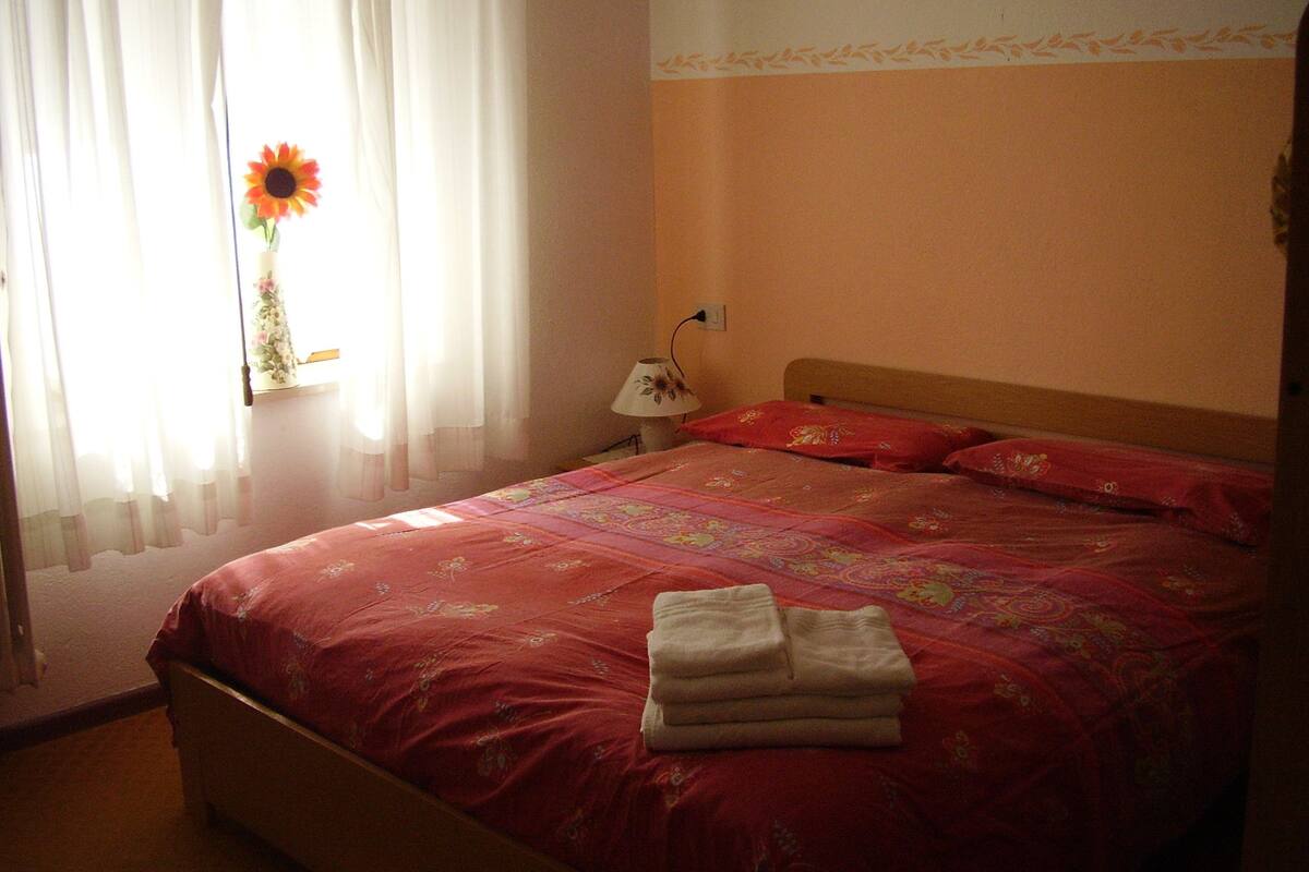 Een net opgemaakt bed met een bloemenbedekker, een lamp op een nachtkastje, en een raam met dunne gordijnen en een zonnebloem.