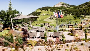 A view of a resort area with outdoor seating, flags, and a mountainous backdrop. - Hotel Continental Dolomites (Selva di Val Gardena)
