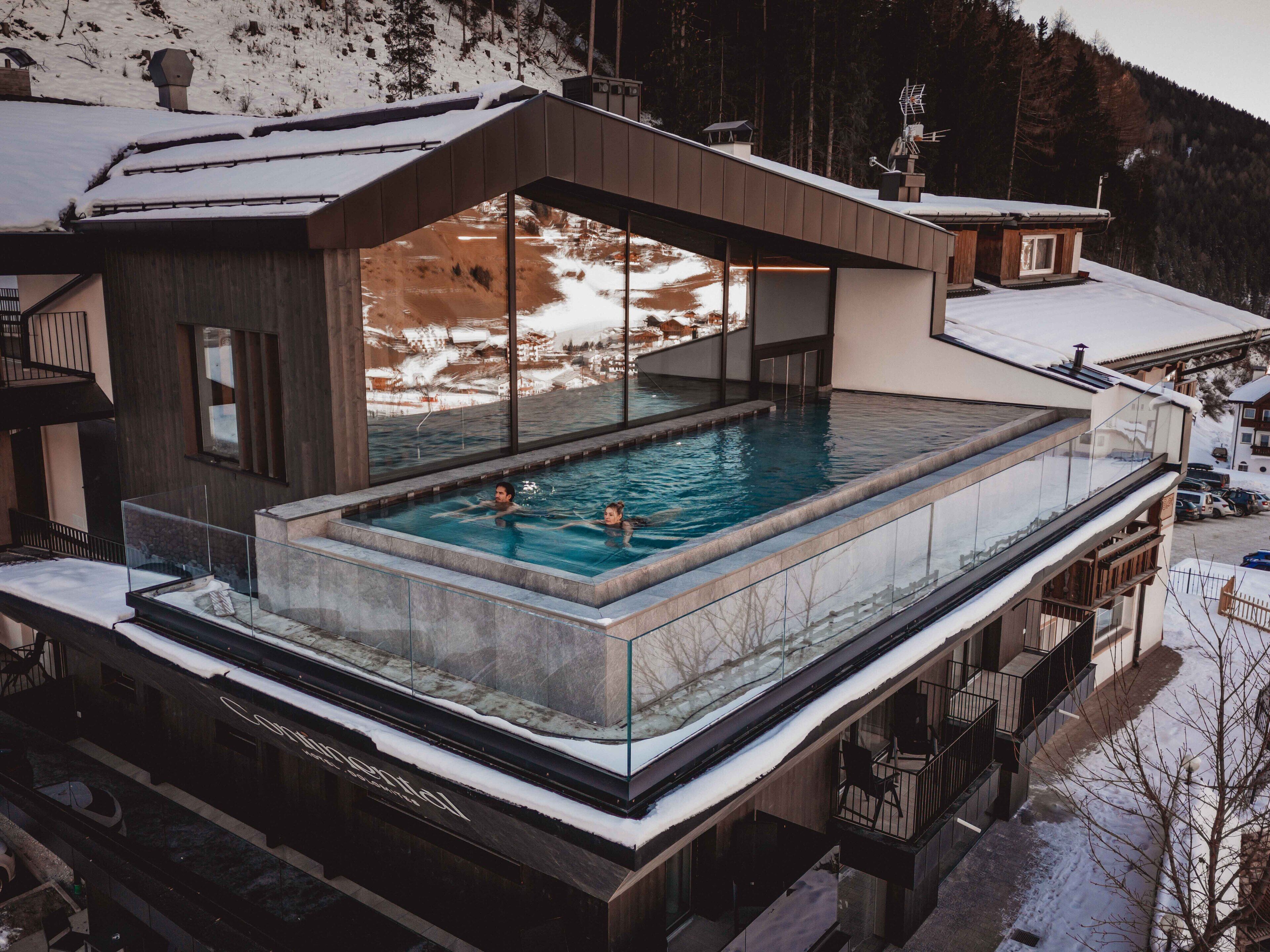 Un edificio moderno con piscina infinita de cristal, vistas a una pista de esquí y fachada de madera.