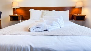 Una cama bien hecha con sábanas blancas y una toalla blanca doblada, flanqueada por dos mesitas de noche con lámparas, en una habitación de hotel.