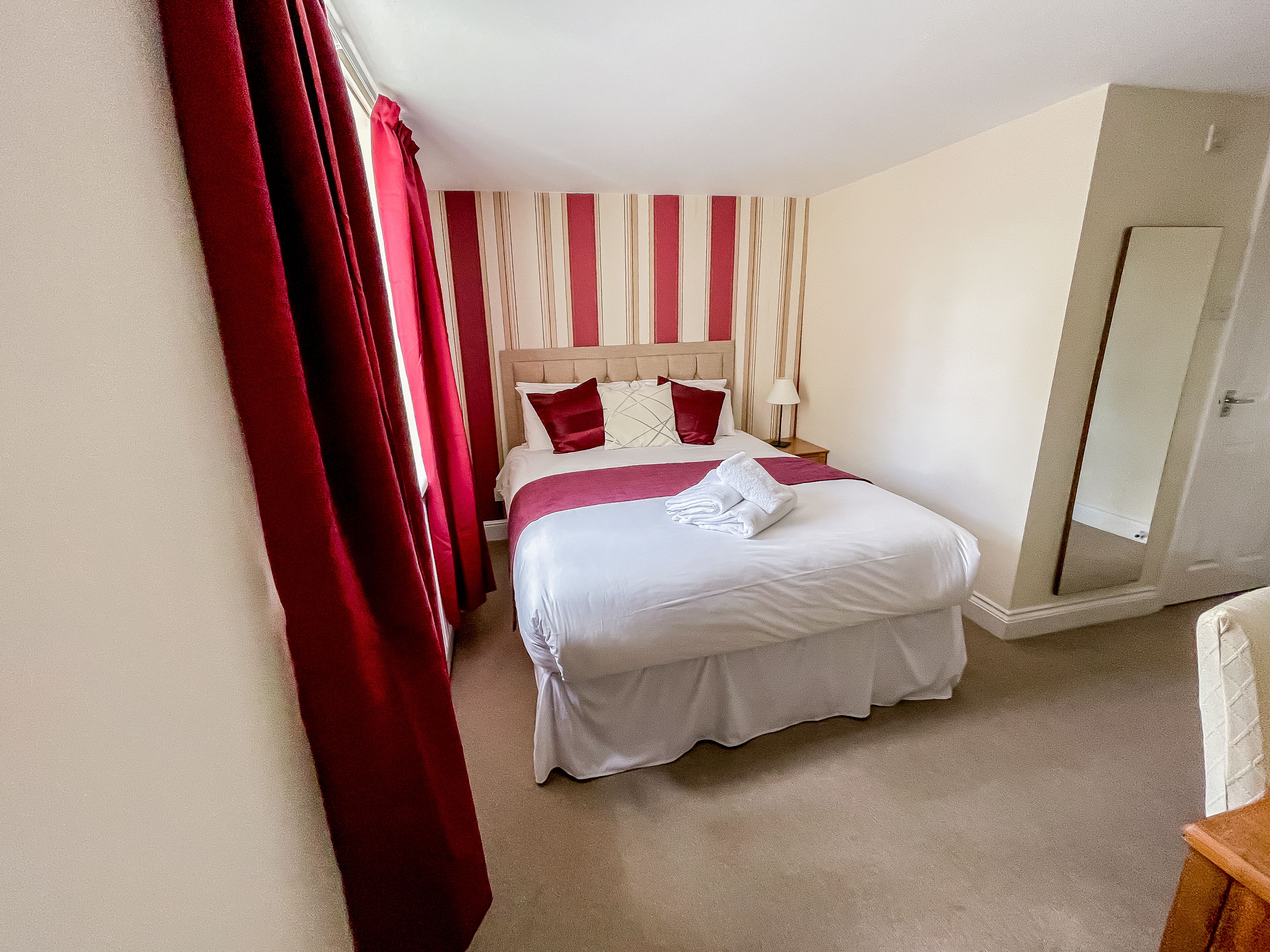 A hotel room with a bed, a nightstand, a lamp, a mirror, and a door.